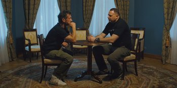 Movie still from “Superpower” (2023), directed by Aaron Kaufman – A couple of men sitting at a wooden table; Medium shot, Over the shoulder angle