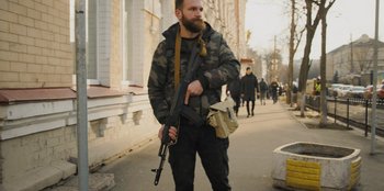Movie still from “Superpower” (2023), directed by Aaron Kaufman – A man with a beard holding a rifle on the sidewalk; Medium shot, Low angle