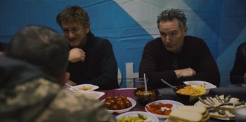 Movie still from “Superpower” (2023), directed by Aaron Kaufman – A couple of men sitting at a table with plates of food on it; Medium shot, Over the shoulder angle
