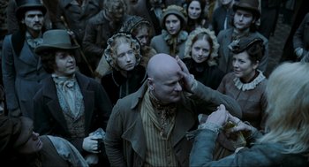 Movie still from “Sweeney Todd: The Demon Barber of Fleet Street” (2007), directed by Tim Burton – A group of people gathered around a bald man; Medium shot, Over the shoulder angle