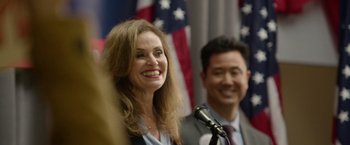 Movie still from “Sweet Girl” (2021), directed by Brian Andrew Mendoza – A woman standing at a podium with a microphone and a man standing next to her; Close Up shot, Over the shoulder angle