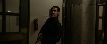 Movie still from “Sweet Girl” (2021), directed by Brian Andrew Mendoza – A man holding a gun while standing next to a wall; Medium shot, Over the shoulder angle