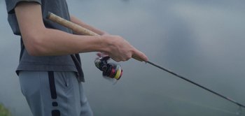 Movie still from “Take Care of Maya” (2023), directed by Henry Roosevelt – A person holding a fishing pole with a fishing reel in it; Extreme Close Up shot, Low angle