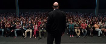 Movie still from “Ted 2” (2015), directed by Seth MacFarlane – A man in a black suit stands in front of an audience; Wide shot, Over the shoulder angle