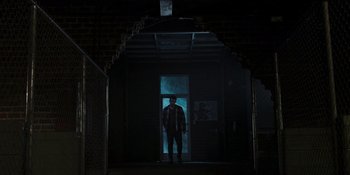 Movie still from “Teen Wolf: The Movie” (2023), directed by Russell Mulcahy – A man standing in a dark room in front of an open door; Wide shot, Low angle