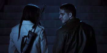 Movie still from “Teen Wolf: The Movie” (2023), directed by Russell Mulcahy – A man and a woman standing next to each other holding guns; Medium shot, Over the shoulder angle