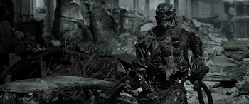Movie still from “Terminator Salvation” (2009), directed by McG – A man with a gun sitting on top of a dirt field; Medium shot, Low angle