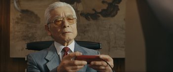 Movie still from “Tetris” (2023), directed by Jon S. Baird – An older man in a suit holding a cell phone in his hands; Close Up shot, Over the shoulder angle