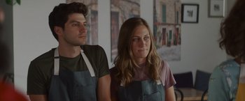 Movie still from “That's Amor” (2022), directed by Shaun Paul Piccinino – A man and a woman wearing aprons standing next to each other; Medium shot, Low angle
