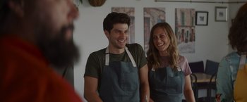 Movie still from “That's Amor” (2022), directed by Shaun Paul Piccinino – A man and a woman wearing aprons smiling for the camera; Medium shot, Over the shoulder angle