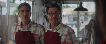 Movie still from “That's Amor” (2022), directed by Shaun Paul Piccinino – A man wearing a red apron and a checkered shirt; Medium shot, Over the shoulder angle
