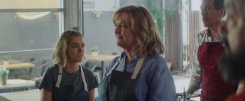 Movie still from “That's Amor” (2022), directed by Shaun Paul Piccinino – A woman standing next to another woman in an apron; Medium shot, Over the shoulder angle