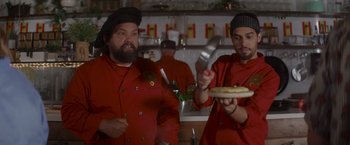Movie still from “That's Amor” (2022), directed by Shaun Paul Piccinino – Two men in chef's outfits holding a pizza and a knife; Medium shot, Low angle