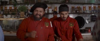 Movie still from “That's Amor” (2022), directed by Shaun Paul Piccinino – Two men in red shirts and black hats in a kitchen; Medium shot, Over the shoulder angle