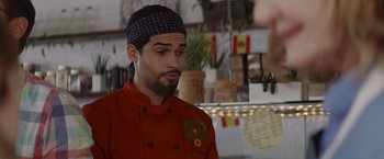 Movie still from “That's Amor” (2022), directed by Shaun Paul Piccinino – A man in a red chef's jacket and a black hat; Close Up shot, Over the shoulder angle