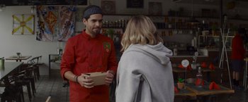 Movie still from “That's Amor” (2022), directed by Shaun Paul Piccinino – A man and a woman are standing in front of a counter; Medium shot, Over the shoulder angle