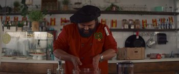 Movie still from “That's Amor” (2022), directed by Shaun Paul Piccinino – A man wearing a red shirt and a black hat is making food; Medium shot, Low angle