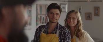 Movie still from “That's Amor” (2022), directed by Shaun Paul Piccinino – A man and a woman wearing aprons posing for a picture; Close Up shot, Over the shoulder angle