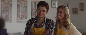Movie still from “That's Amor” (2022), directed by Shaun Paul Piccinino – A man and a woman wearing yellow aprons smiling; Close Up shot, Over the shoulder angle