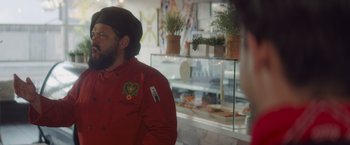 Movie still from “That's Amor” (2022), directed by Shaun Paul Piccinino – A man wearing a red jacket in front of a display case; Medium shot, Over the shoulder angle