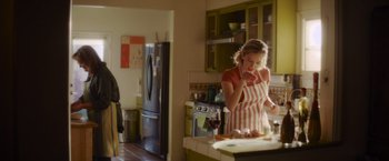 Movie still from “That's Amor” (2022), directed by Shaun Paul Piccinino – A woman in an apron standing in a kitchen talking on the phone; Medium shot, Over the shoulder angle
