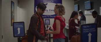 Movie still from “That's Amor” (2022), directed by Shaun Paul Piccinino – A man and a woman standing next to each other in front of an airport sign; Medium shot, Over the shoulder angle