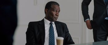 Movie still from “The 355” (2022), directed by Simon Kinberg – A man in a suit and tie sitting in front of a cup; Close Up shot, Over the shoulder angle