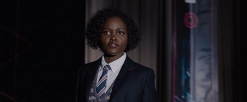Movie still from “The 355” (2022), directed by Simon Kinberg – A woman wearing a suit and tie standing in front of a curtain; Medium shot, Over the shoulder angle