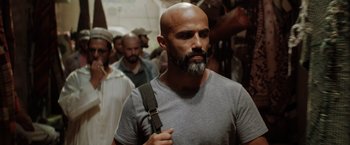 Movie still from “The 355” (2022), directed by Simon Kinberg – A man with a beard is holding a bag in front of a group of people; Close Up shot, Over the shoulder angle