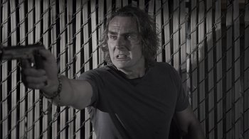 Movie still from “The Amityville Moon” (2021), directed by Thomas J. Churchill – A man in a black t - shirt is standing in front of a chain link fence; Close Up shot, Low angle