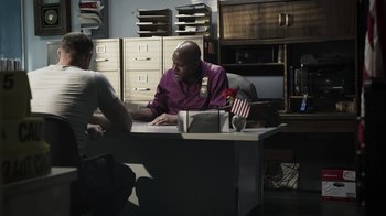 Movie still from “The Amityville Moon” (2021), directed by Thomas J. Churchill – A man sitting at a desk in front of a computer with an american flag; Medium shot, Over the shoulder angle