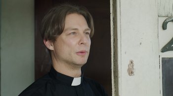 Movie still from “The Amityville Moon” (2021), directed by Thomas J. Churchill – A man wearing a priest's outfit looking to his left; Close Up shot, Low angle