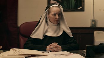 Movie still from “The Amityville Moon” (2021), directed by Thomas J. Churchill – A woman sitting at a desk with her hands clasped together; Medium shot, Low angle