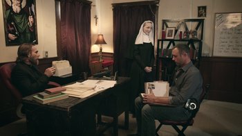 Movie still from “The Amityville Moon” (2021), directed by Thomas J. Churchill – A nun standing next to a man in a room; Wide shot, High angle