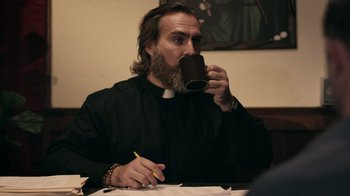 Movie still from “The Amityville Moon” (2021), directed by Thomas J. Churchill – A priest drinking a cup of coffee while sitting at a table; Medium shot, Over the shoulder angle