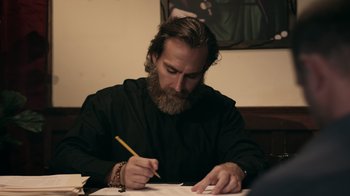 Movie still from “The Amityville Moon” (2021), directed by Thomas J. Churchill – A man with a beard writing on a piece of paper with a pencil; Medium shot, Over the shoulder angle