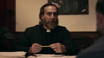 Movie still from “The Amityville Moon” (2021), directed by Thomas J. Churchill – A man with a long beard and a priest's collar is sitting at a table with a pencil; Close Up shot, Over the shoulder angle