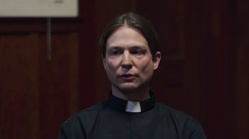 Movie still from “The Amityville Moon” (2021), directed by Thomas J. Churchill – A man wearing a priest's collar and standing in front of a wooden wall; Close Up shot, Low angle