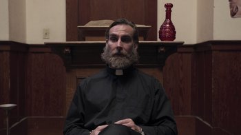 Movie still from “The Amityville Moon” (2021), directed by Thomas J. Churchill – A man sitting in front of a table with a vase; Close Up shot, Low angle