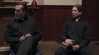 Movie still from “The Amityville Moon” (2021), directed by Thomas J. Churchill – Two men in black robes sitting in a room; Medium shot, Over the shoulder angle