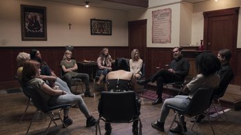 Movie still from “The Amityville Moon” (2021), directed by Thomas J. Churchill – A group of people sitting in a circle in a room; Wide shot, High angle
