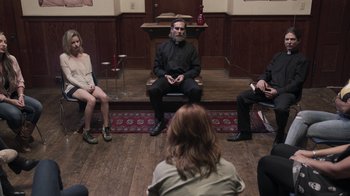 Movie still from “The Amityville Moon” (2021), directed by Thomas J. Churchill – A group of people sitting in a circle in front of a woman; Wide shot, High angle