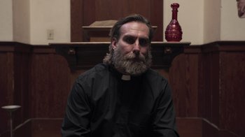 Movie still from “The Amityville Moon” (2021), directed by Thomas J. Churchill – A man with long hair and a beard wearing a priest's outfit; Close Up shot, Low angle
