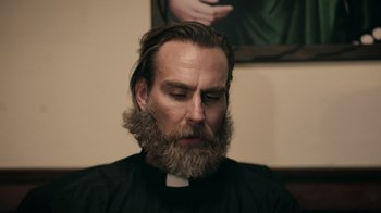 Movie still from “The Amityville Moon” (2021), directed by Thomas J. Churchill – A man with long hair and a beard is wearing a priest's outfit; Close Up shot, Low angle