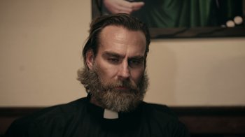 Movie still from “The Amityville Moon” (2021), directed by Thomas J. Churchill – A man with long hair and a beard wearing a priest's outfit; Close Up shot, Low angle