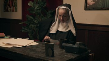 Movie still from “The Amityville Moon” (2021), directed by Thomas J. Churchill – A nun sitting at a table looking at papers; Medium shot, Low angle