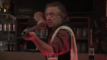 Movie still from “The Amityville Moon” (2021), directed by Thomas J. Churchill – An older man is holding a wooden stick and a glass of beer; Close Up shot, Low angle