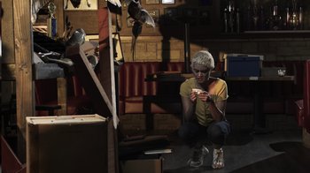 Movie still from “The Amityville Moon” (2021), directed by Thomas J. Churchill – A woman sitting on the ground holding a cell phone; Wide shot, Low angle