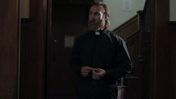 Movie still from “The Amityville Moon” (2021), directed by Thomas J. Churchill – A man with a long beard wearing a black shirt; Medium shot, Low angle