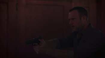 Movie still from “The Amityville Moon” (2021), directed by Thomas J. Churchill – A man holding a gun in a dark room; Medium shot, Over the shoulder angle